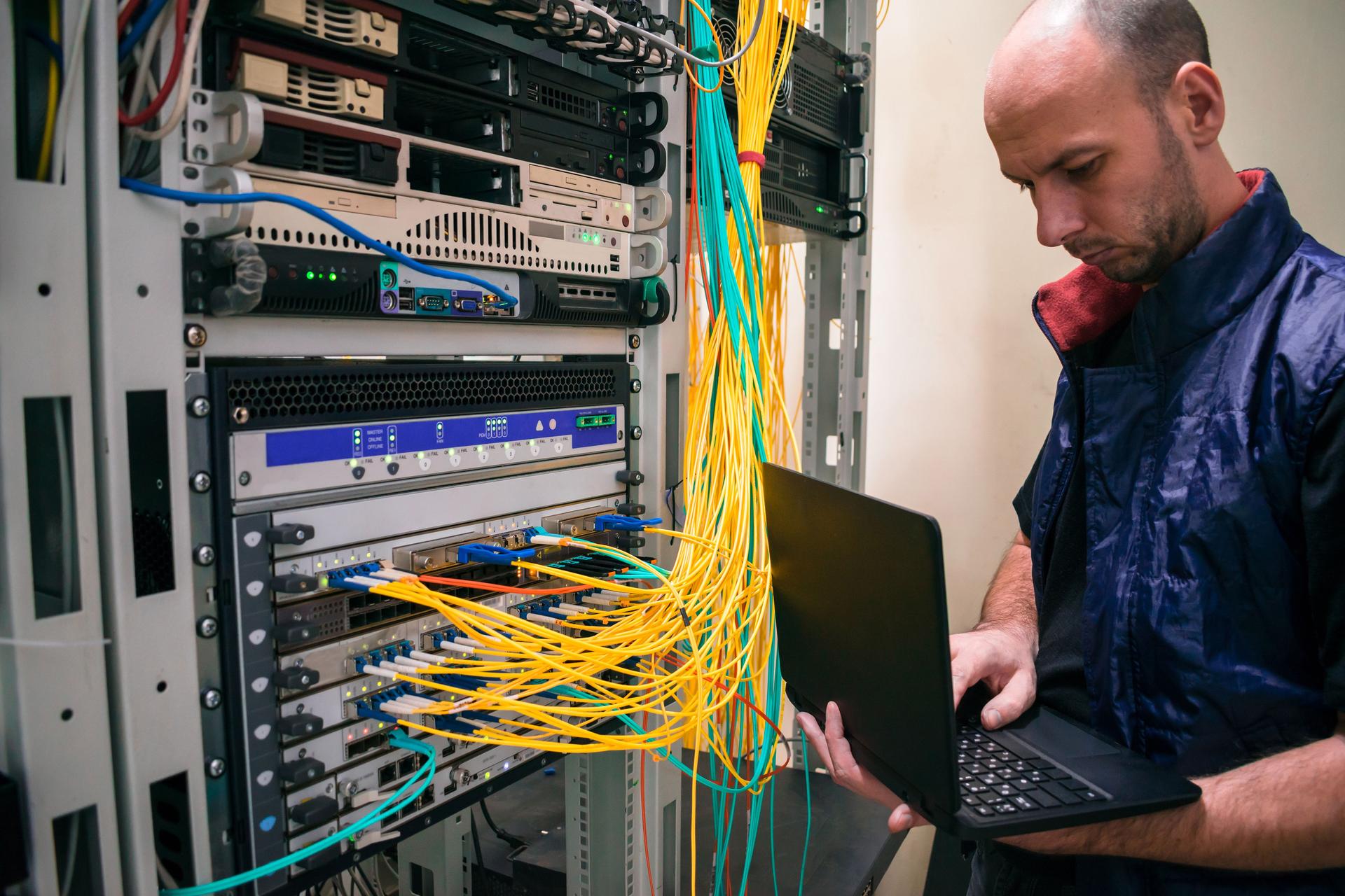 The system administrator with a laptop is in the server room. A man sets up a central router in a datacenter. Configuring server hardware.